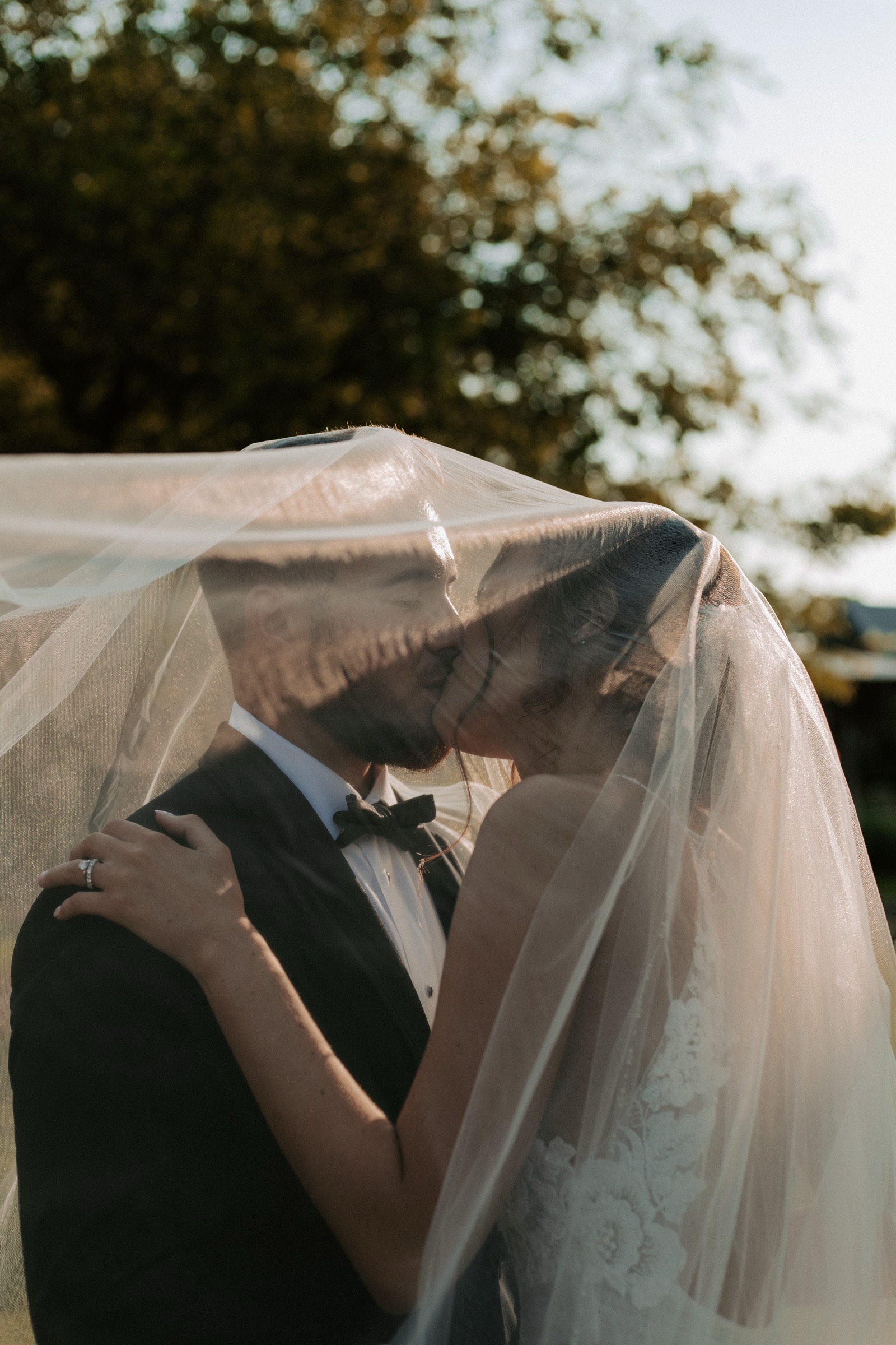 Bride and groom kissing under a veil, backlit by trees at golden hour