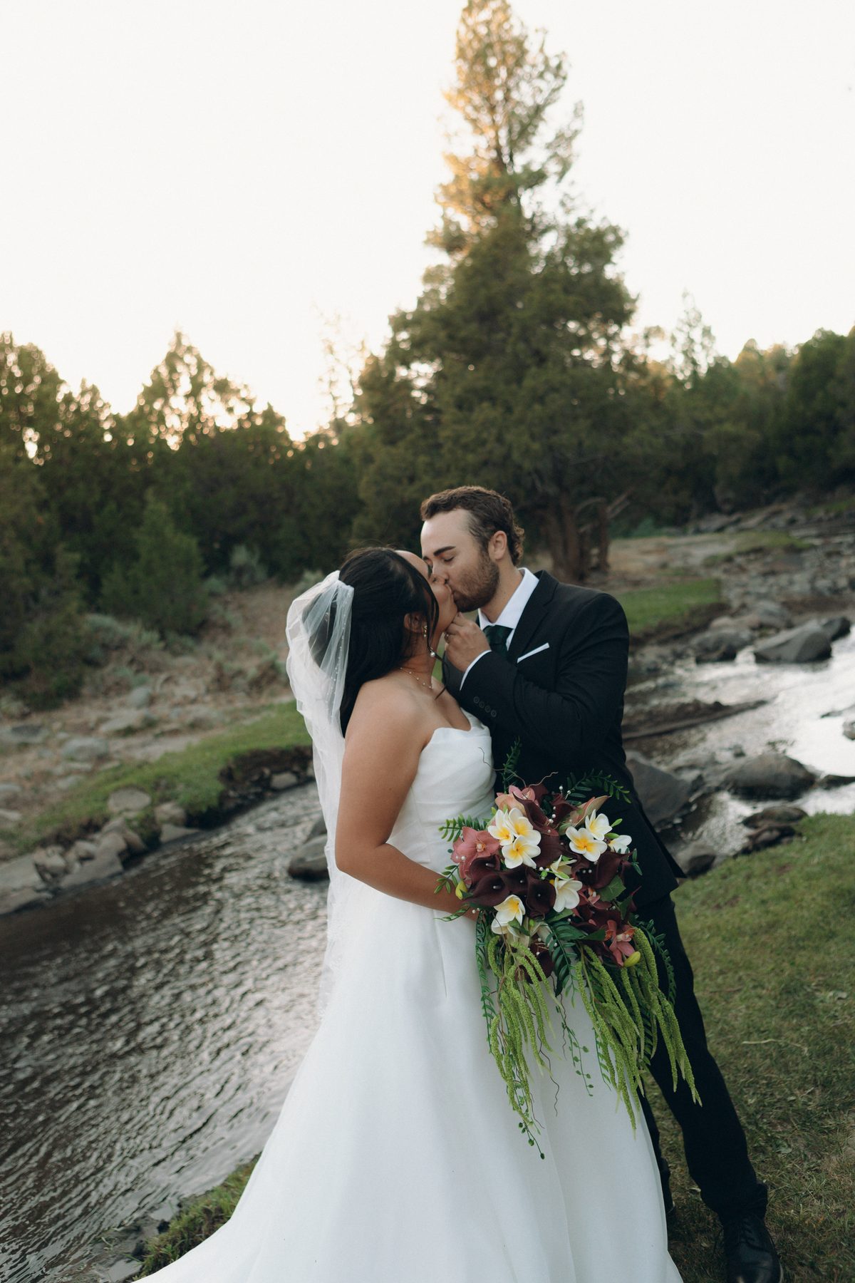 Bride and groom embracing by a river, wedding veil catching the breeze